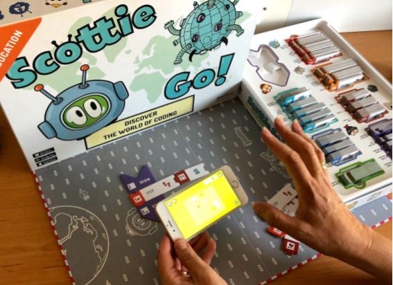 A child's hands holding a phone, scanning a line of Scottie Go programming tiles laid out on the game board